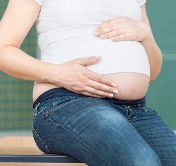 Midsection,Of,Pregnant,Teacher,Sitting,On,Desk,In,Classroom